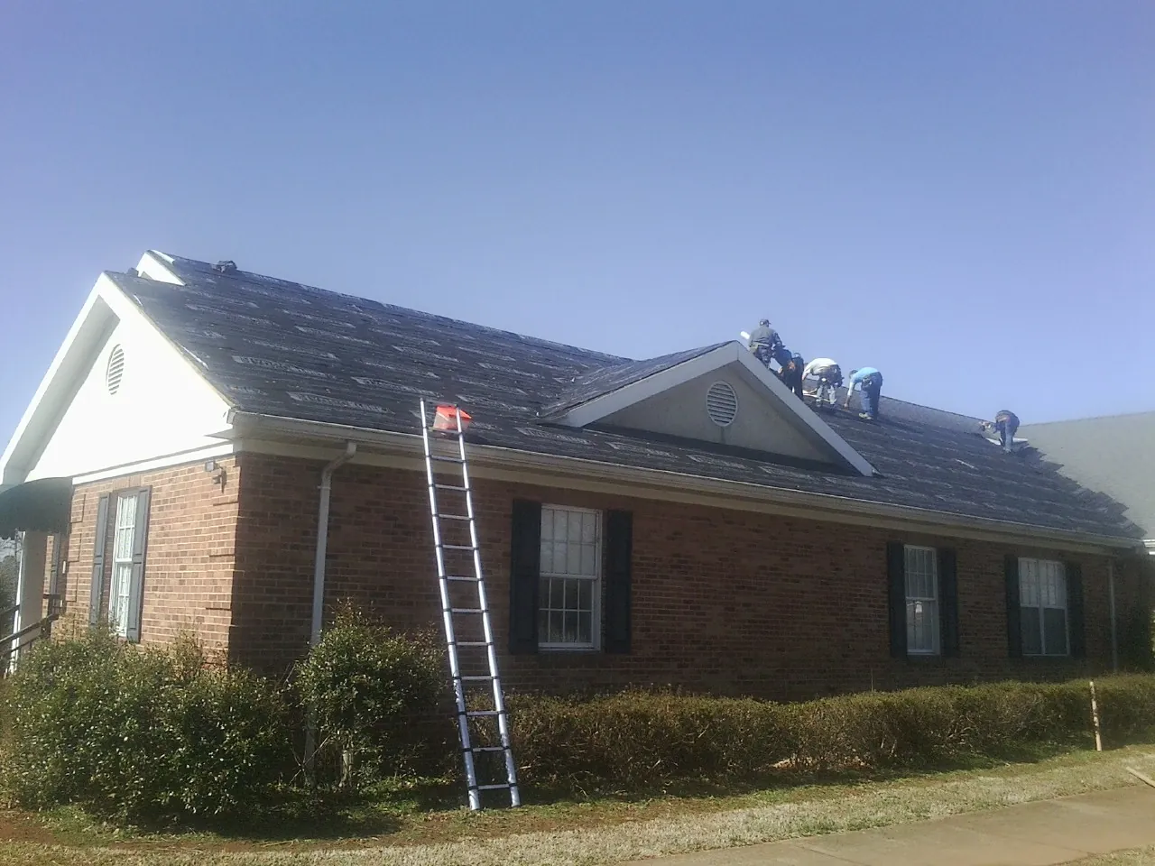 Crew tearing shingles off a brick ranch roof with a ladder set — Metal Roof Repair work by our crew in Glasford