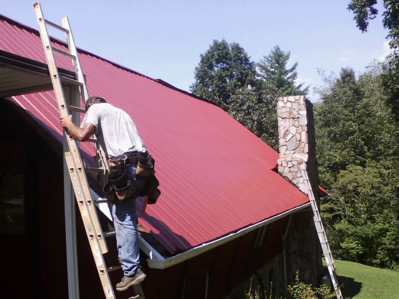 Roofer on a ladder beside a red metal roof and stone chimney — Soffit & Fascia Repair by Roofing Brothers in Glasford